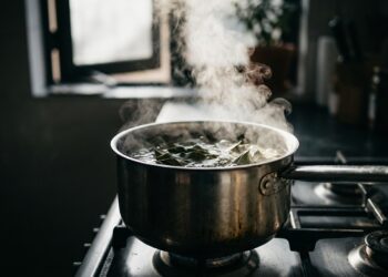 Panela de inox com folhas de louro e vinagre branco fervendo no fogão, com vapor subindo e janela da cozinha ao fundo