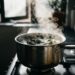 Panela de inox com folhas de louro e vinagre branco fervendo no fogão, com vapor subindo e janela da cozinha ao fundo
