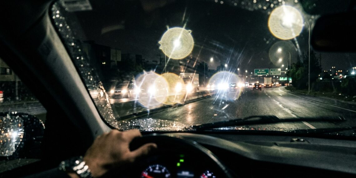 Visão do motorista através do para-brisa à noite com faróis de veículos formando halos luminosos e ofuscamento na pista molhada.