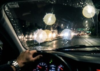 Visão do motorista através do para-brisa à noite com faróis de veículos formando halos luminosos e ofuscamento na pista molhada.