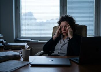 Profissional sentado em mesa de escritório com expressão de exaustão profunda e mãos na cabeça sob luz matinal suave