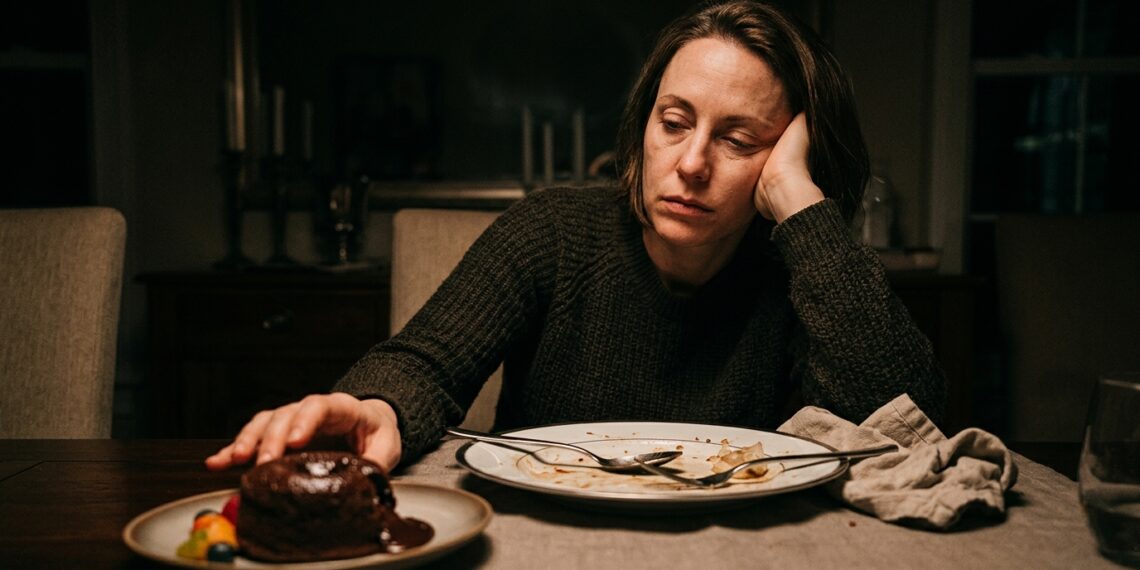 Pessoa com expressão de cansaço segurando a cabeça após o almoço diante de uma mesa de refeição