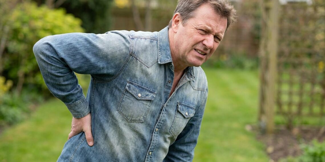 Homem de meia-idade vestindo camisa jeans com expressão de dor, segurando a região lombar das costas em um ambiente externo arborizado.
