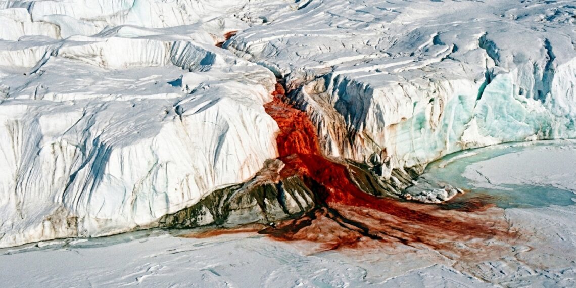 Cachoeira de água vermelho-ferrugem desaguando entre fendas e paredões de gelo branco em uma geleira na Antártida.
