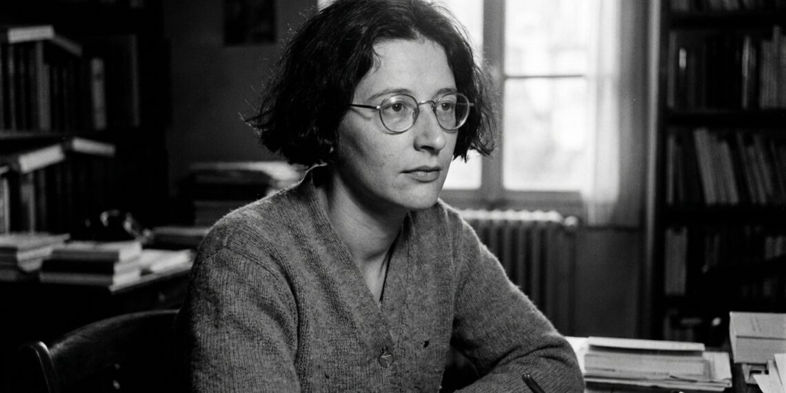 Retrato em preto e branco da filósofa francesa Simone Weil usando óculos de armação redonda e suéter, sentada em uma mesa de estudos com livros ao fundo