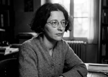 Retrato em preto e branco da filósofa francesa Simone Weil usando óculos de armação redonda e suéter, sentada em uma mesa de estudos com livros ao fundo