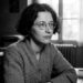 Retrato em preto e branco da filósofa francesa Simone Weil usando óculos de armação redonda e suéter, sentada em uma mesa de estudos com livros ao fundo