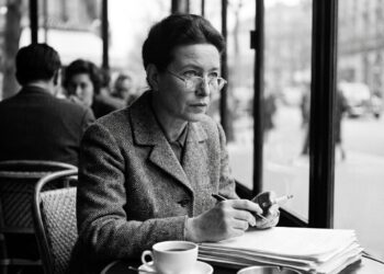 Uma fotografia em preto e branco de Simone de Beauvoir sentada em uma mesa de bistrô em um café parisiense, perto de uma janela