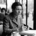 Uma fotografia em preto e branco de Simone de Beauvoir sentada em uma mesa de bistrô em um café parisiense, perto de uma janela
