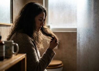 Mulher em um banheiro iluminado segurando as pontas do cabelo opaco e ressecado com expressão de preocupação.