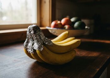 Cacho de bananas maduras com o talo envolto em papel-alumínio sobre bancada de madeira, com fruteira de maçãs e abacates ao fundo desfocado.