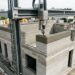 Impressora 3D de grande porte deposita material cimentício nas paredes de um prédio em construção.