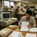 Profissionais trabalham com documentos impressos, telefone fixo e computador de tubo em escritório do fim dos anos 1980.