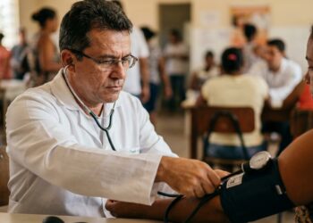 Médico aferindo a pressão arterial do braço de um paciente com estetoscópio e esfigmomanômetro em posto de saúde.