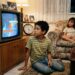 Criança espera diante de uma televisão de tubo pelo início de um programa em uma sala típica dos anos 1990.