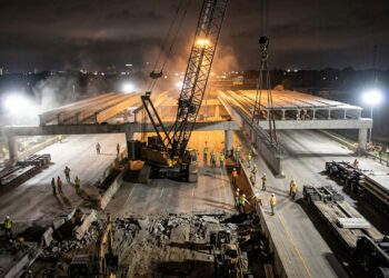 Guindaste instala uma viga pré-fabricada durante a substituição acelerada de uma ponte sobre uma rodovia.