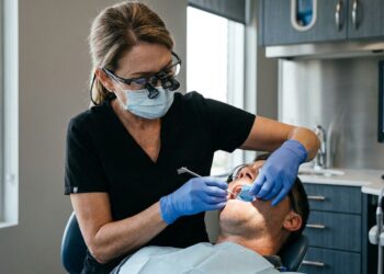 Periodontista com lupa cirúrgica e luvas examinando a mordida e a estabilidade dental de paciente na cadeira odontológica.