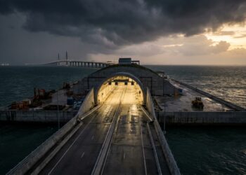 Vista aérea panorâmica da travessia marítima mostrando uma ponte estaiada que se conecta a uma ilha artificial oval e mergulha em direção a um túnel submarino no mar aberto.