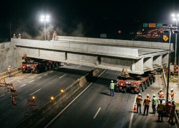 Grande trecho pré-fabricado de ponte é transportado sobre carretas SPMT para instalação acima de uma rodovia.