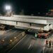 Grande trecho pré-fabricado de ponte é transportado sobre carretas SPMT para instalação acima de uma rodovia.