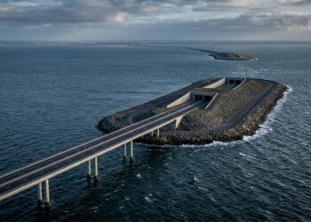 Rodovia sobre viadutos de concreto cruzando o mar e descendo em rampa rumo ao portal de um túnel submarino em uma ilha artificial de pedras