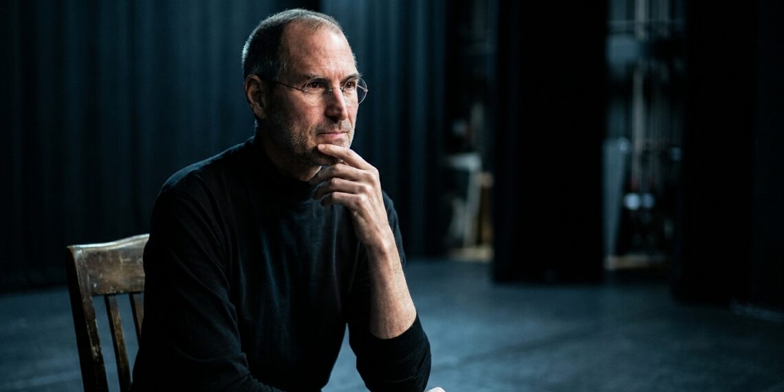 Steve Jobs de perfil reflexivo em palco escuro com iluminação pontual suave