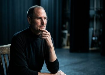 Steve Jobs de perfil reflexivo em palco escuro com iluminação pontual suave