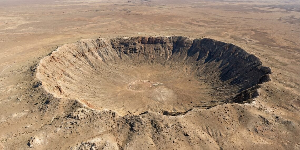 Vista aérea em ângulo aberto de uma grande cratera de impacto de meteorito em meio a um platô desértico árido.
