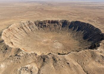 Vista aérea em ângulo aberto de uma grande cratera de impacto de meteorito em meio a um platô desértico árido.