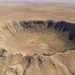 Vista aérea em ângulo aberto de uma grande cratera de impacto de meteorito em meio a um platô desértico árido.