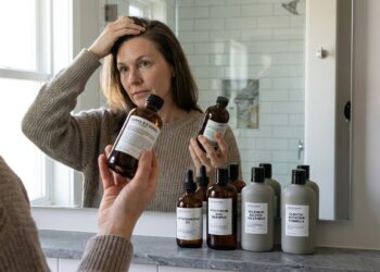 Pessoa examinando a raiz do cabelo no espelho do banheiro com frascos de shampoo dermatológico sobre a bancada.