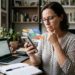 Mulher interrompe uma tarefa ao olhar notificações no celular enquanto trabalha diante de uma mesa.