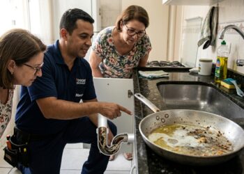 Encanador examina tubulação de pia com acúmulo de gordura enquanto uma frigideira suja permanece próxima ao ralo.