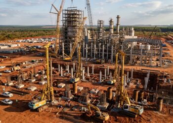 Perfuratrizes executam estacas profundas no solo avermelhado diante de grandes estruturas de uma fábrica de celulose em construção.