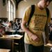 Estudante com camiseta chamativa caminhando em sala de aula enquanto outros alunos continuam concentrados em cadernos