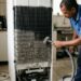 Técnico limpa a serpentina traseira coberta de poeira de uma geladeira.