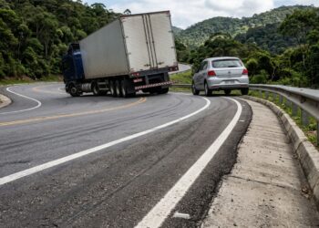 Caminhão e carro percorrem uma curva onde o pavimento sobe em direção ao lado externo.