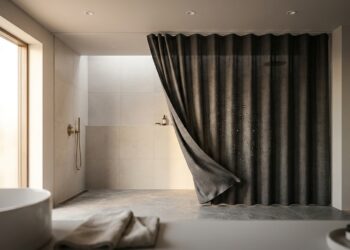 Cortina impermeável moderna com gotas de água perladas em sua superfície, pendurada em trilho aéreo discreto em banheiro contemporâneo com piso limpo