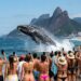 Baleia-franca saltando no mar diante de banhistas que filmam a cena com celulares na praia do Leblon, com o Morro Dois Irmãos ao fundo.