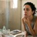 Mulher adulta com sinais de cansaço observa lesões de acne no queixo e na mandíbula diante do espelho.
