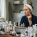 Mulher observa vermelhidão e pequenas espinhas no rosto diante de bancada cheia de produtos de skincare.