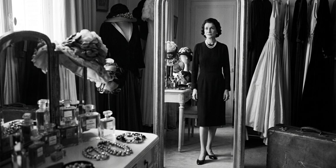 Coco Chanel diante do espelho em um ambiente luxuoso, usando um visual simples e elegante.