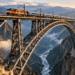Ponte ferroviária metálica com arco de aço atravessando um vale profundo entre montanhas, com um trem sobre a estrutura.