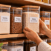 Potes transparentes com tampas de bambu e etiquetas de identificação organizados em prateleiras, contendo arroz, feijão, farinha, macarrão e outros mantimentos.