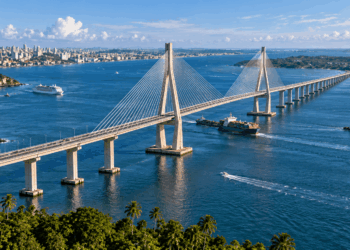Vista aérea ilustrativa de uma grande ponte estaiada atravessando a Baía de Todos-os-Santos, com navios, águas azuis e áreas urbanas costeiras.