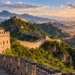 Grande Muralha da China atravessando montanhas e regiões áridas, com torres de vigilância e muralhas de pedra ao nascer do sol.