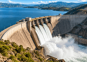 Barragem monumental de concreto retendo um grande reservatório em um vale montanhoso, com vertedouros liberando água em grande volume.