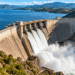 Barragem monumental de concreto retendo um grande reservatório em um vale montanhoso, com vertedouros liberando água em grande volume.