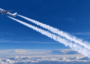 Avião comercial voando acima das nuvens e deixando dois rastros brancos no céu azul.
