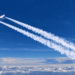 Avião comercial voando acima das nuvens e deixando dois rastros brancos no céu azul.